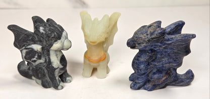 Three small dragon figurines made of natural stone, displayed side by side on a white surface with a light gray marble-patterned background. The left figurine is black and white marbled, the center figurine is pale cream with a small orange accent, and the right figurine is deep blue with natural stone texture.