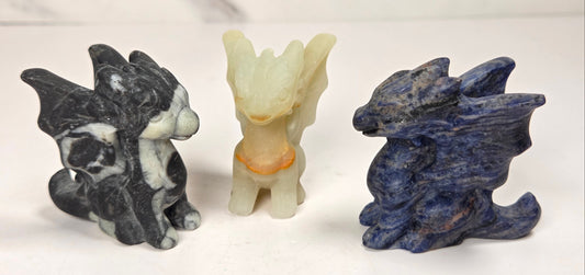 Three small dragon figurines made of natural stone, displayed side by side on a white surface with a light gray marble-patterned background. The left figurine is black and white marbled, the center figurine is pale cream with a small orange accent, and the right figurine is deep blue with natural stone texture.