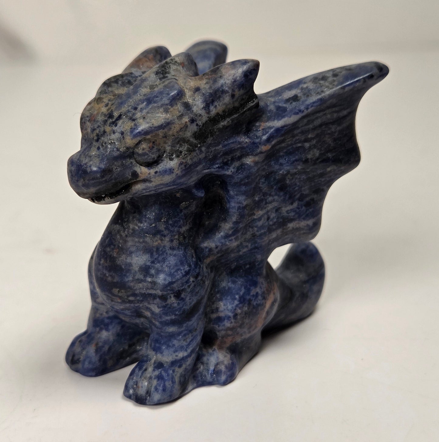 A small blue stone dragon figurine with textured lapis lazuli-like coloring, standing on a plain light surface against a light gray background.