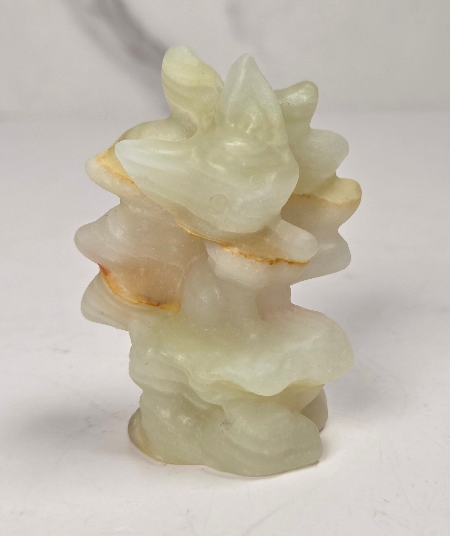 A small carved stone figurine of a stylized animal, likely a rabbit or deer, made from pale greenish-white jade or similar material with light brown natural inclusions, set against a white marble-patterned background.