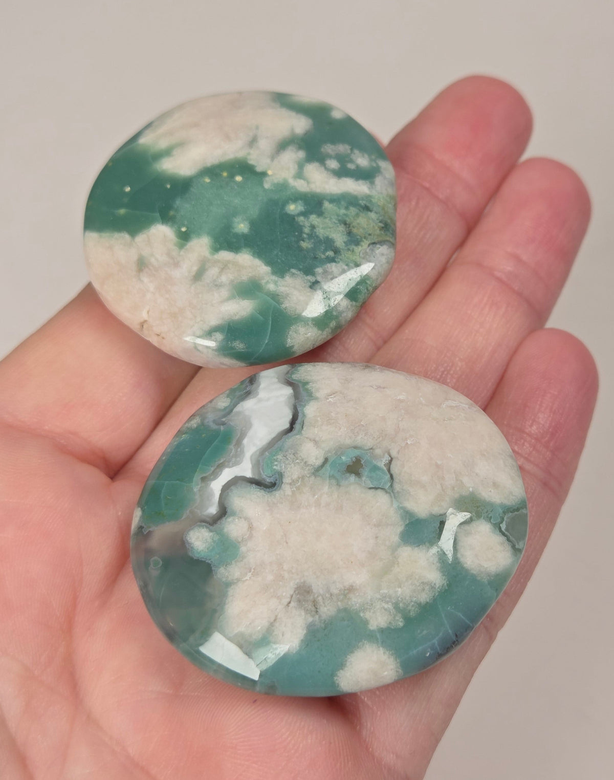 Two polished oval gemstones with a green and white marbled pattern, held in a person's hand against a plain light background.