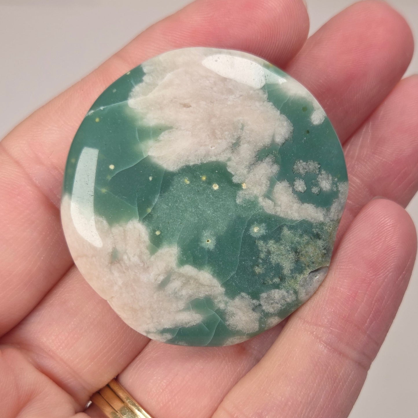 A polished round stone with a green and white marbled pattern, held in a person's hand. The stone has a glossy finish and is shown close-up against a soft, light background.