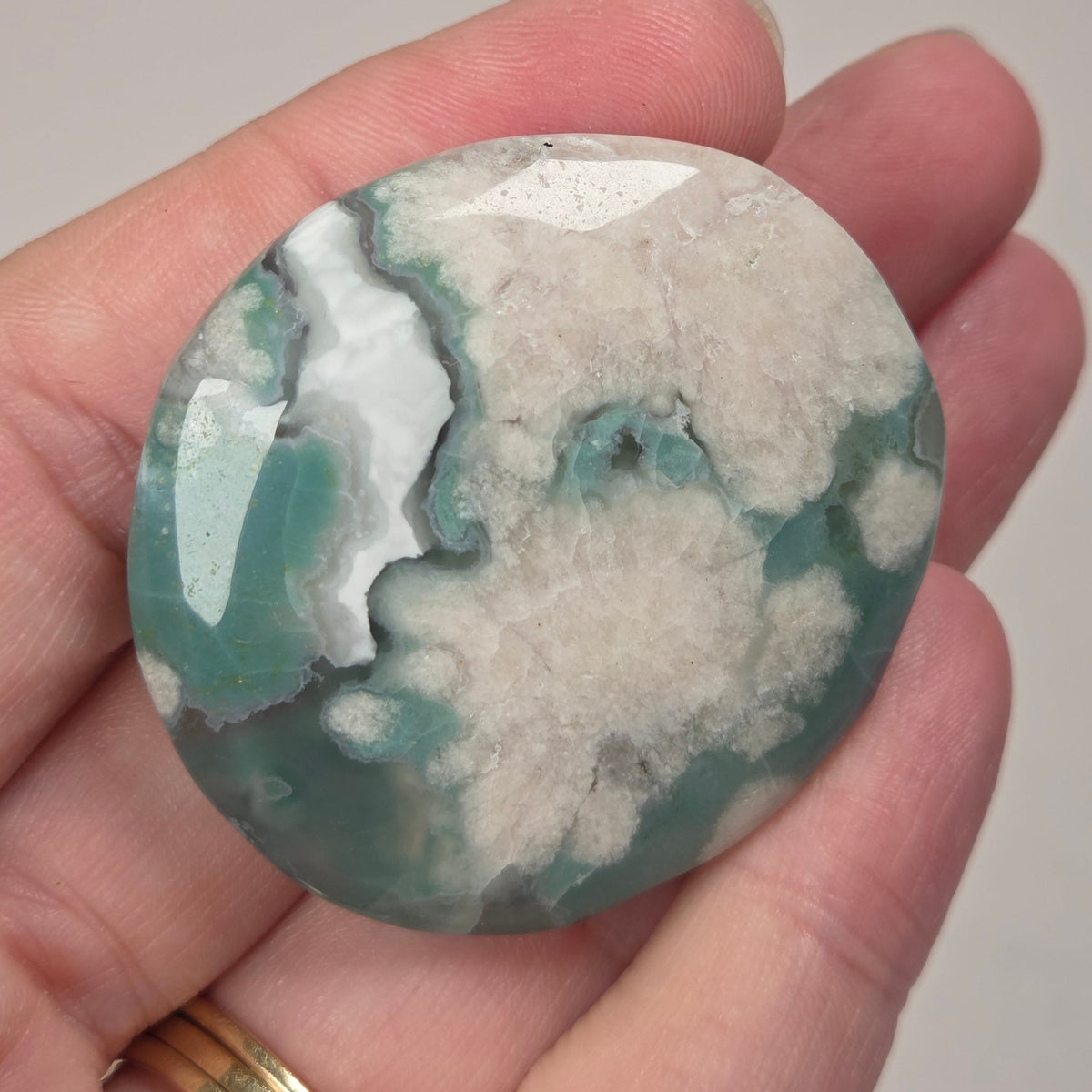 A polished round stone with a mottled pattern of teal green, white, and gray, held in a person's hand. The hand is resting against a light gray marble-patterned background.