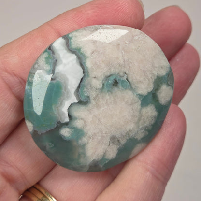 A polished round stone with a mottled pattern of teal green, white, and gray, held in a person's hand. The hand is resting against a light gray marble-patterned background.