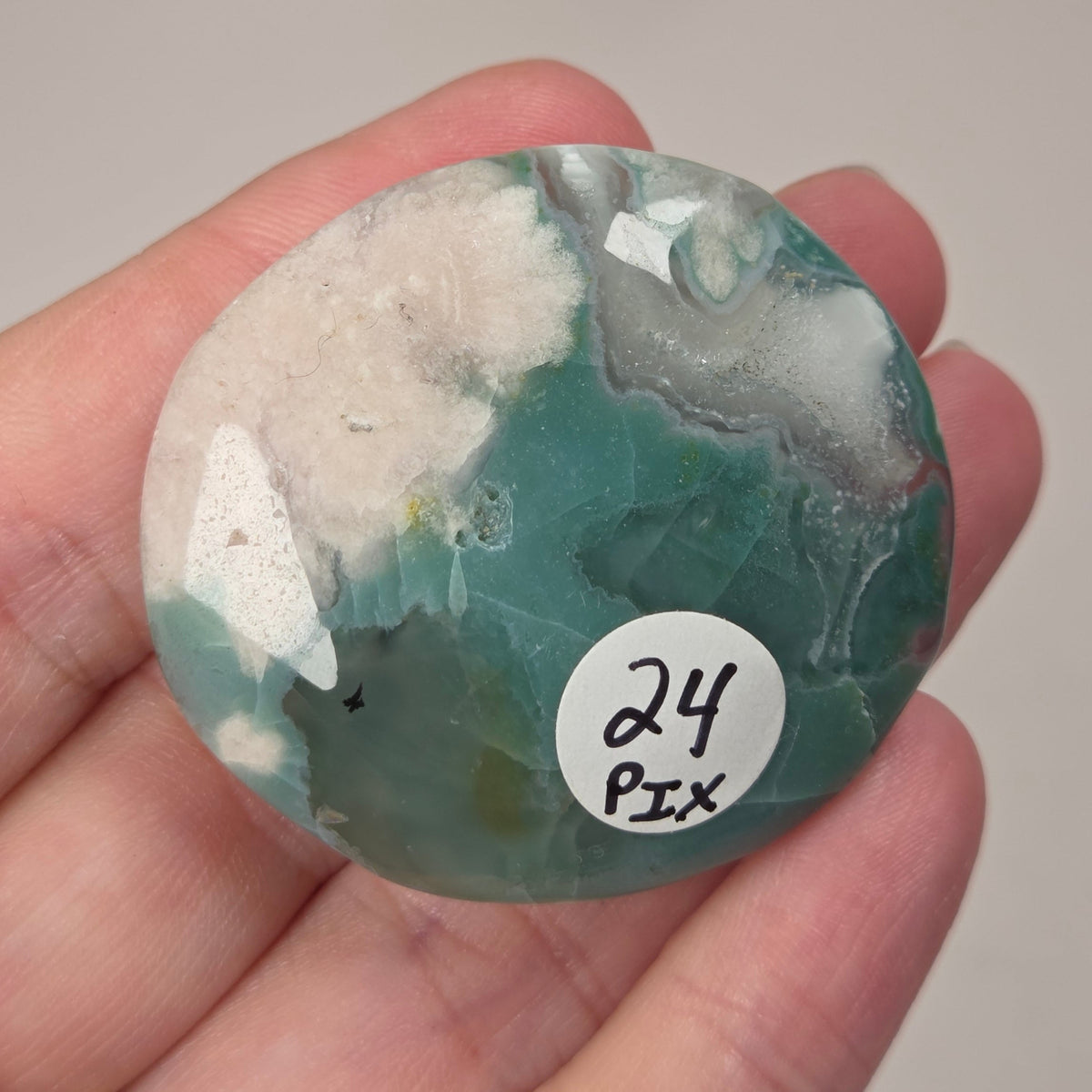 A hand holds a polished round stone with a marbled pattern of teal green, white, and gray. A small white sticker on the stone reads '24 Pix'.