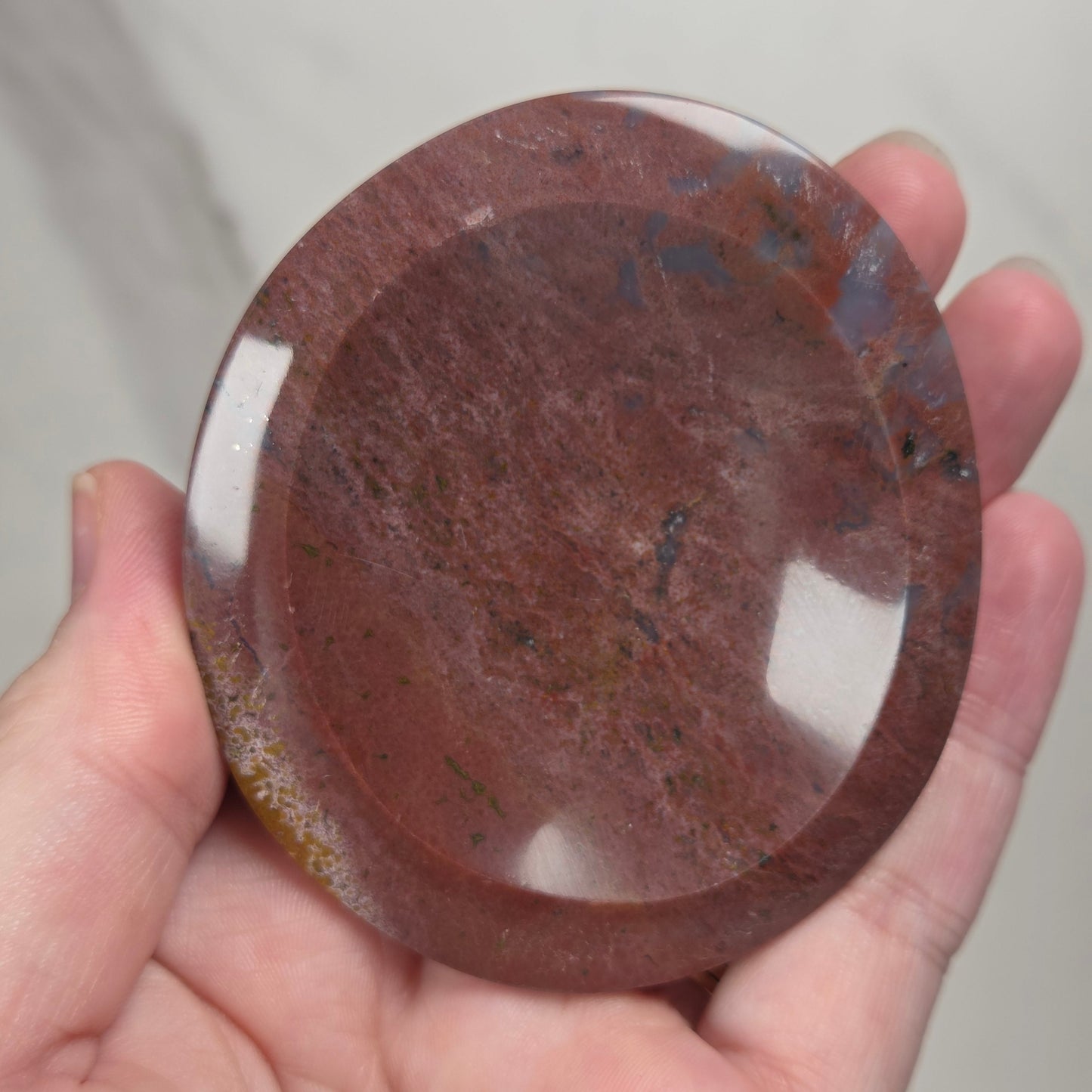 A hand holding a round, polished reddish-brown stone or glass plate with a glossy finish and natural marbled texture.