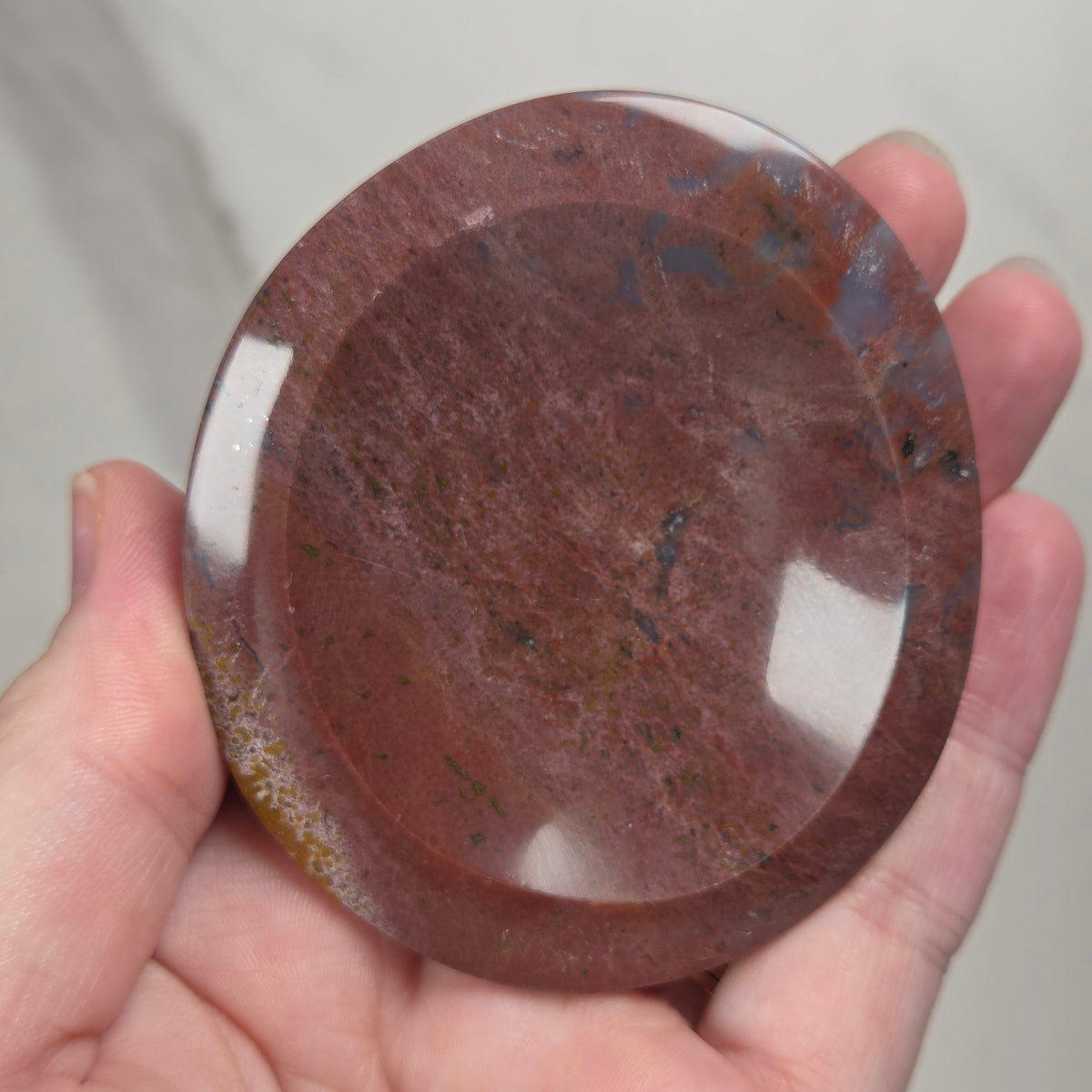 A hand holding a round, polished reddish-brown stone or glass plate with a glossy finish and natural marbled texture.