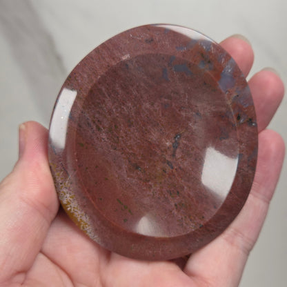 A hand holding a round, polished reddish-brown stone or glass plate with a glossy finish and natural marbled texture.