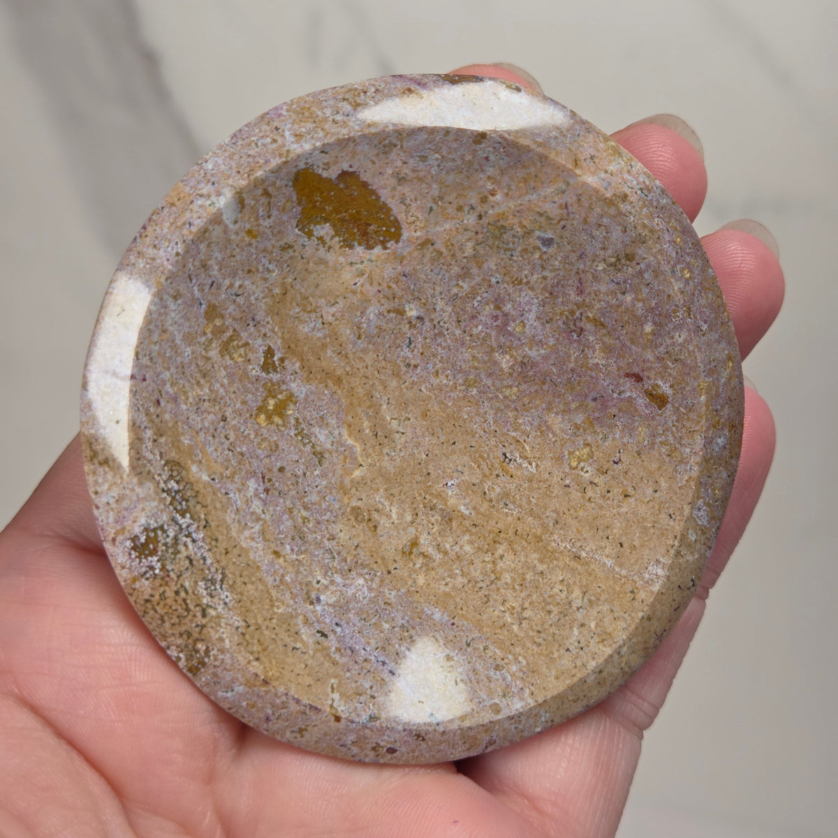A hand holding a round polished stone or mineral slab with natural marbled patterns in beige, brown, gray, and small purple tones, against a light marble-like background.
