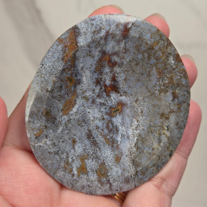 A hand holding a round, polished stone with a mottled gray, white, and rust-brown marbled pattern against a light marble background.