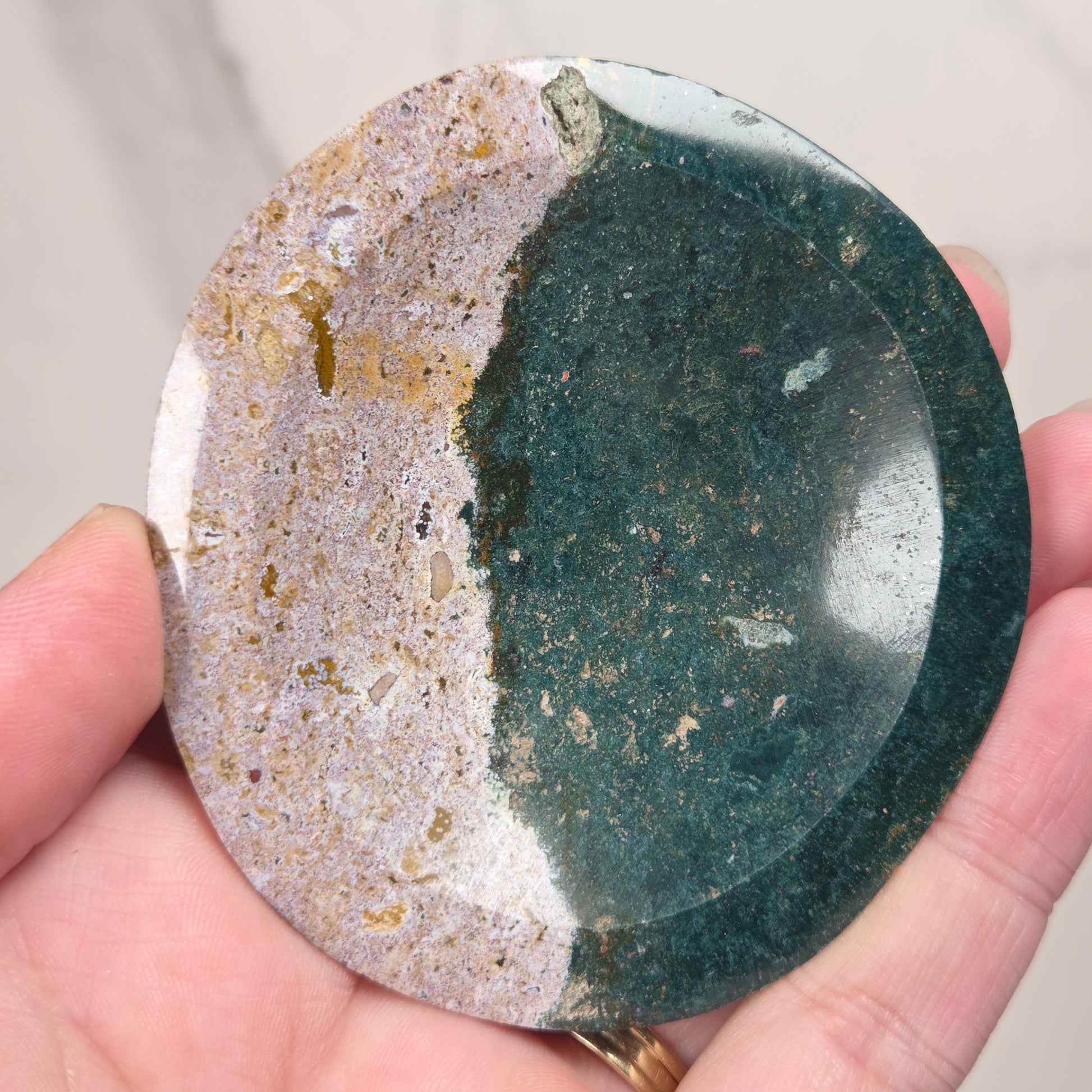 A hand holding a round polished stone with a split design, featuring a dark green textured half and a beige and white marbled half.