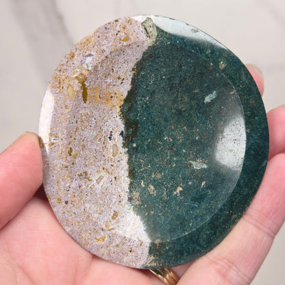 A hand holding a round polished stone with a split design, featuring a dark green textured half and a beige and white marbled half.