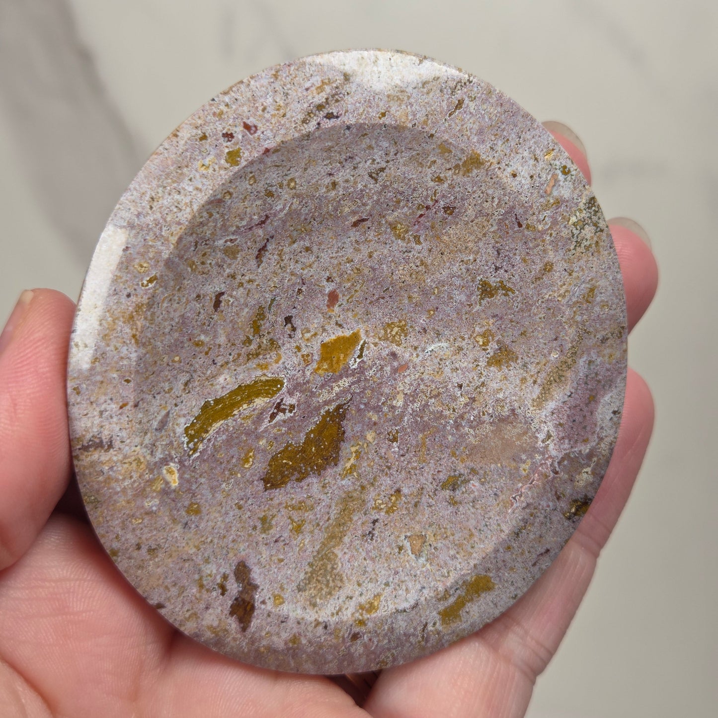 A hand holding a round, shallow stone dish or tray with a speckled marbled pattern in gray, beige, and gold tones.