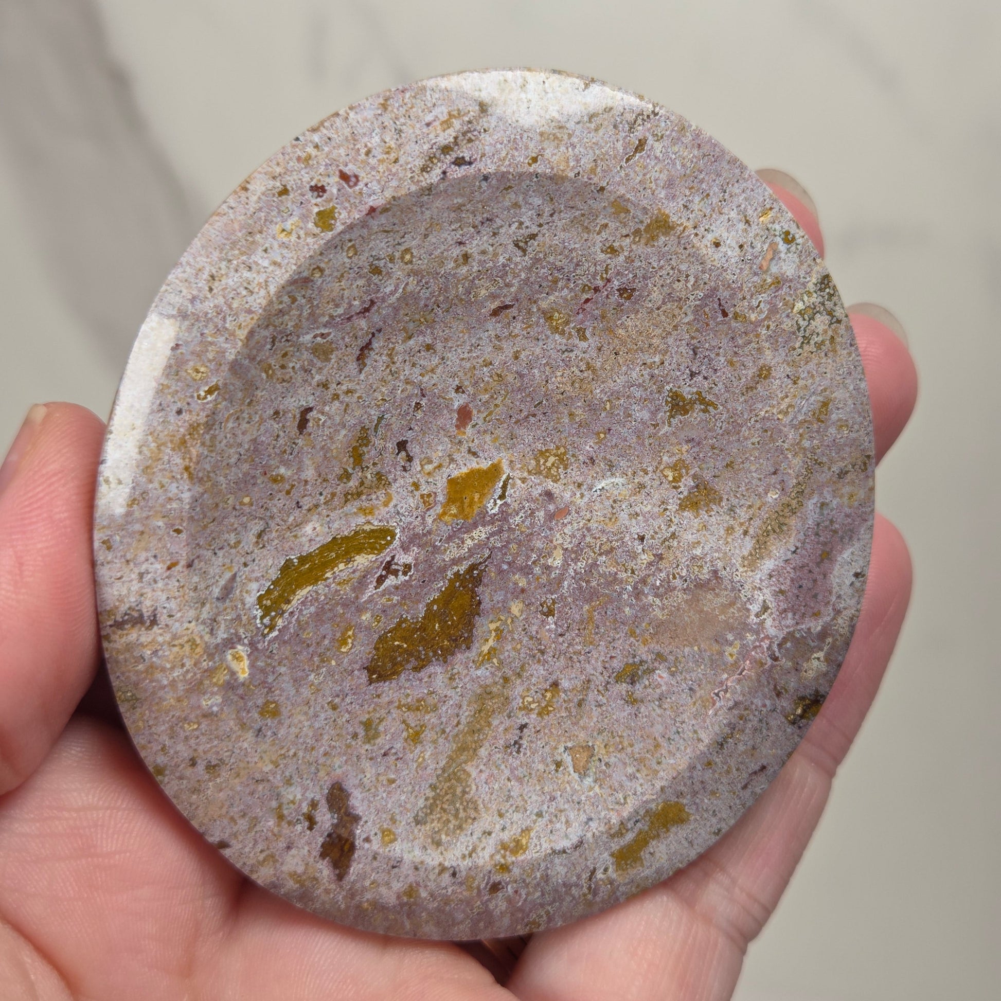A hand holding a round, shallow stone dish or tray with a speckled marbled pattern in gray, beige, and gold tones.