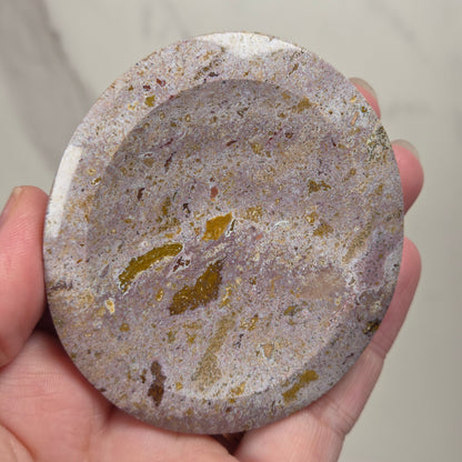 A hand holding a round, shallow stone dish or tray with a speckled marbled pattern in gray, beige, and gold tones.