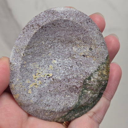 A hand holding a round, textured stone or ceramic dish with a speckled gray and purple surface and small yellow-green areas, against a light gray marble background.