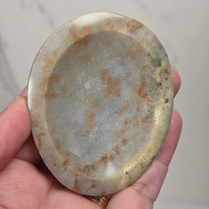 A hand holding an oval-shaped polished stone or jade pendant with natural earthy brown and gray marbling, set against a light gray marble background.