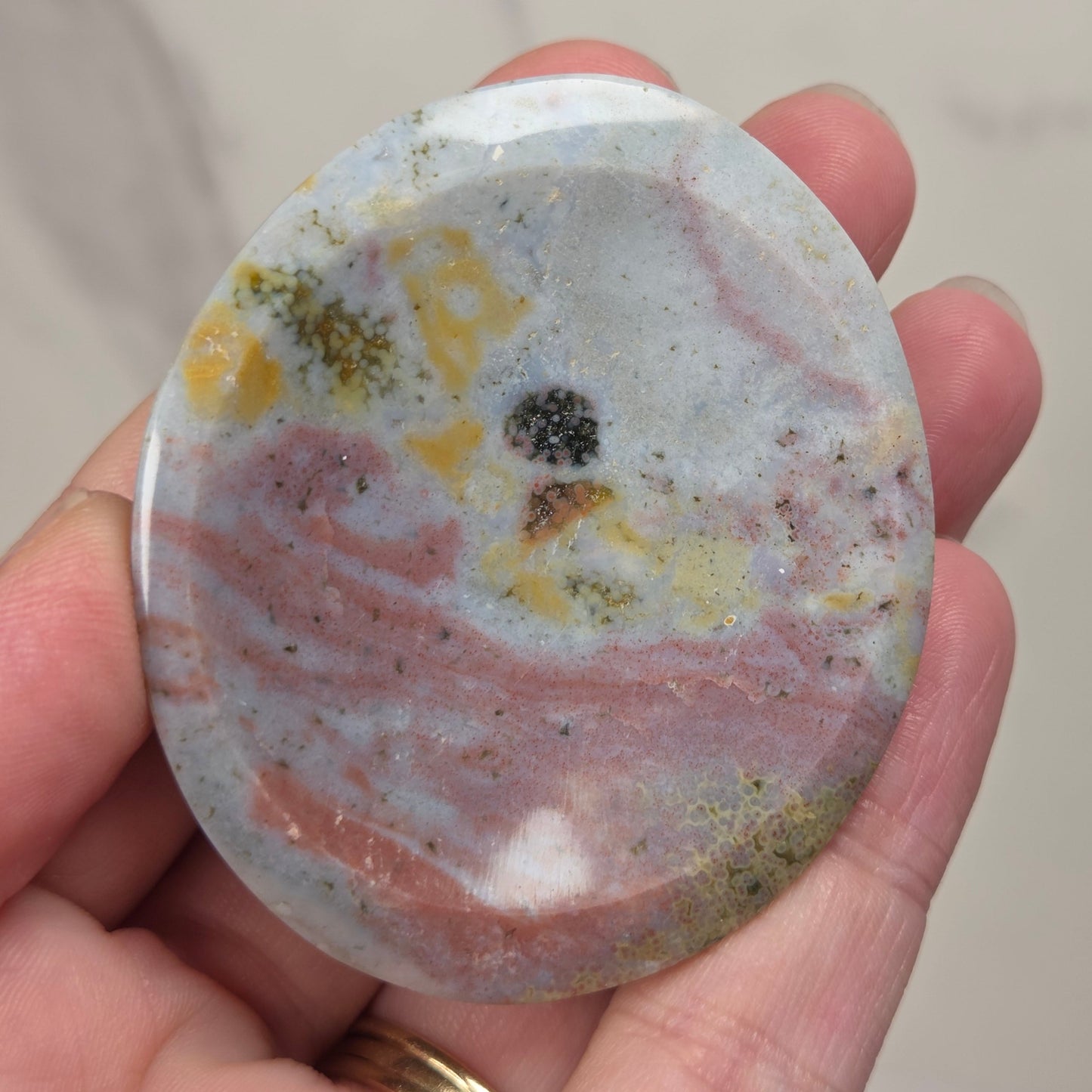 A hand holding a polished oval stone cabochon with marbled pink, white, gray, yellow, and black patterns, set against a light marble background.