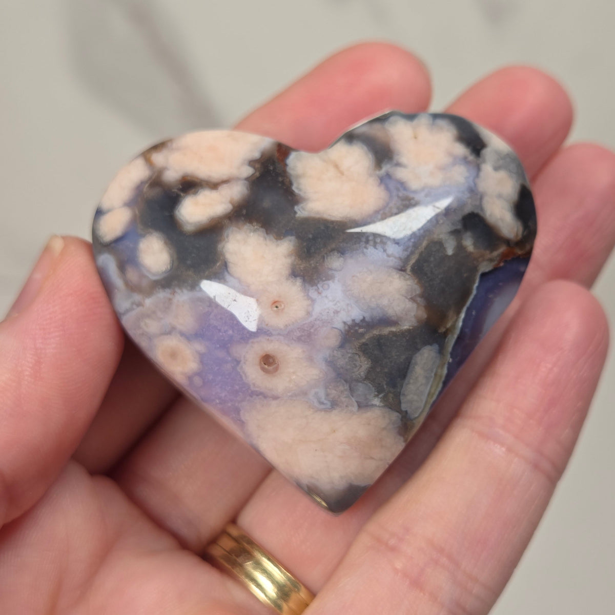 A heart-shaped polished stone with a marbled pattern of beige, purple, gray, and black, held in a person's hand.