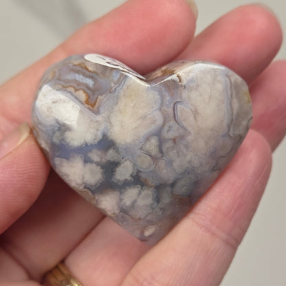 A heart-shaped polished agate stone with natural marbled patterns of gray, white, and brown, held in a person's hand.