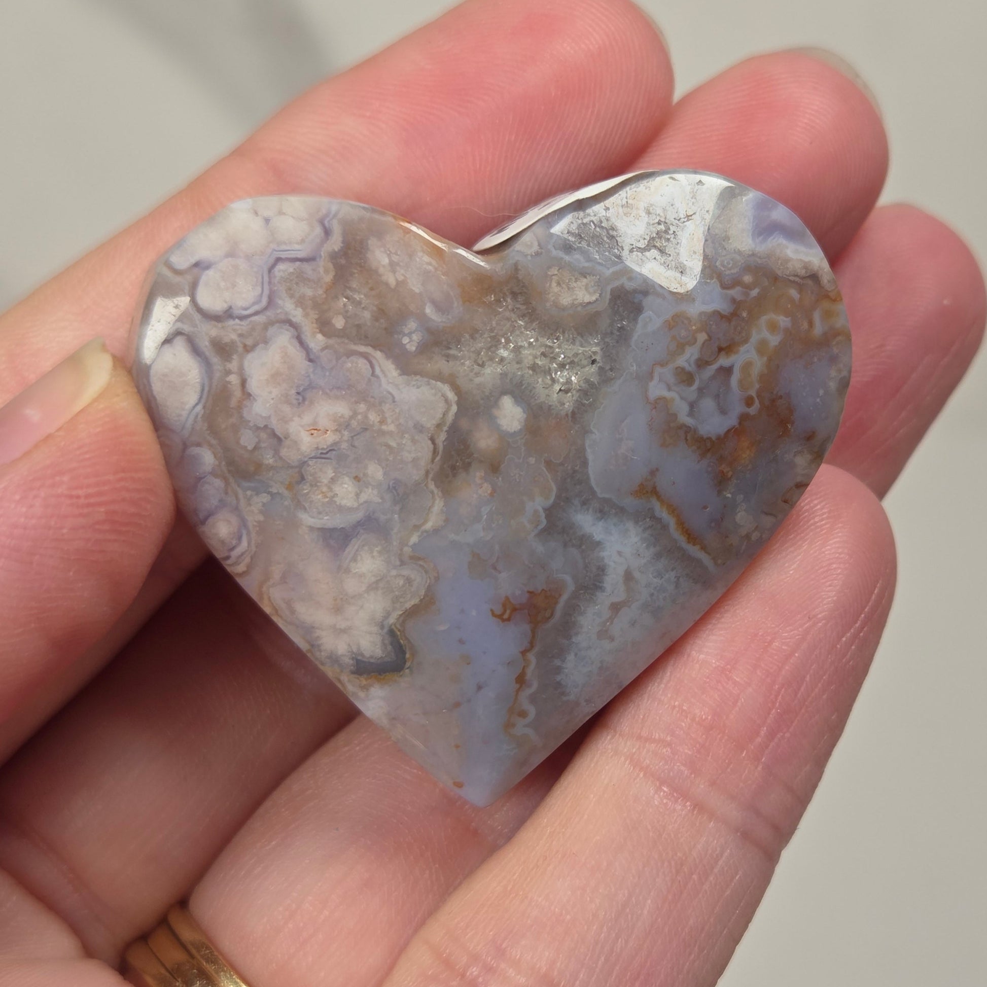 A heart-shaped polished stone pendant with a mottled pattern of gray, white, and brown, held in a person's hand. The pendant has a small silver bail at the top for hanging on a necklace.