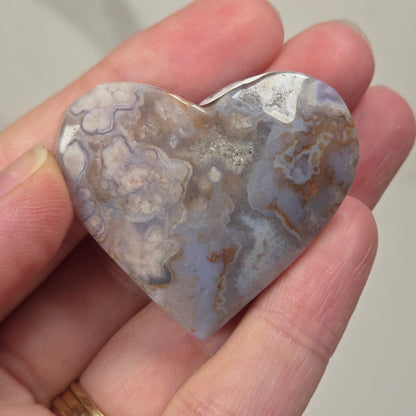 A heart-shaped polished stone pendant with a mottled pattern of gray, white, and brown, held in a person's hand. The pendant has a small silver bail at the top for hanging on a necklace.