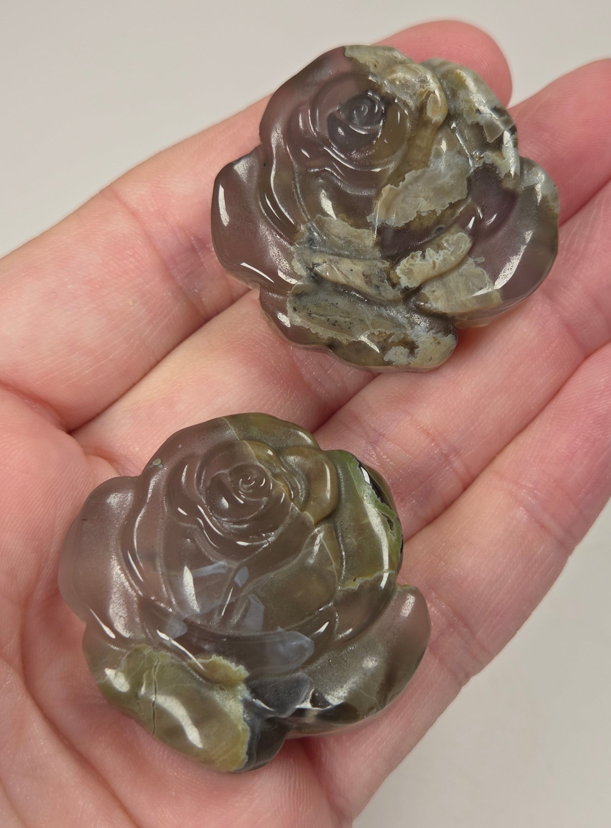 Two carved rose-shaped stone ornaments with a mottled gray, green, and brown pattern, held in a person's hand against a plain light background.
