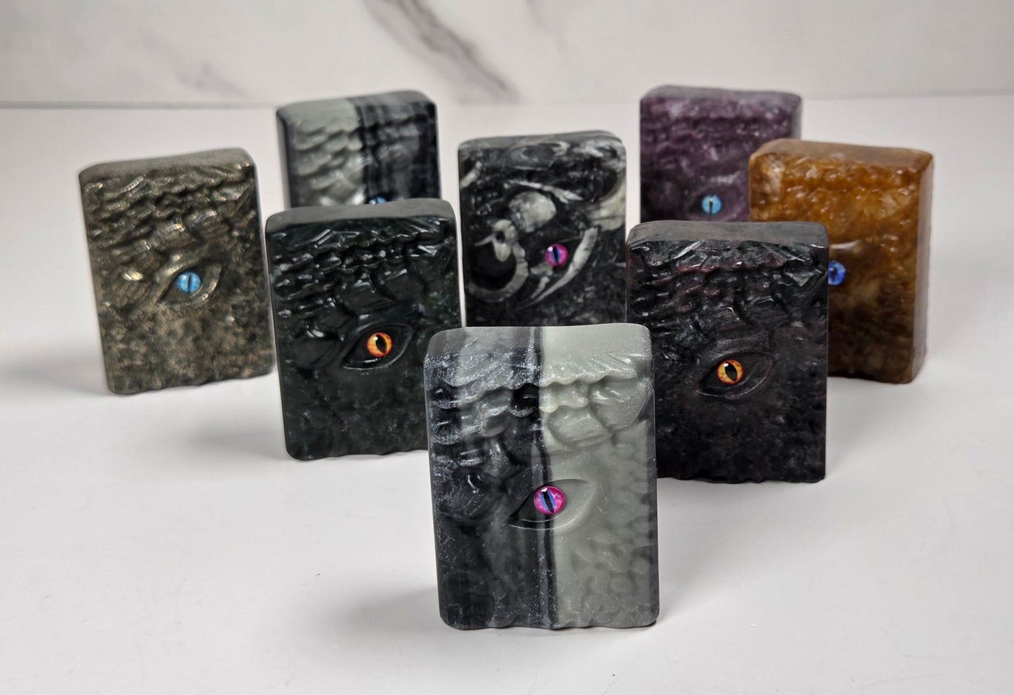 A set of nine small rectangular stone or resin blocks with a textured, organic marbled surface, each featuring a tiny colorful gem or iridescent eye detail. The set includes various colors and patterns such as black, gray, purple, brown, and green, arranged on a white surface with a white marble-patterned background.