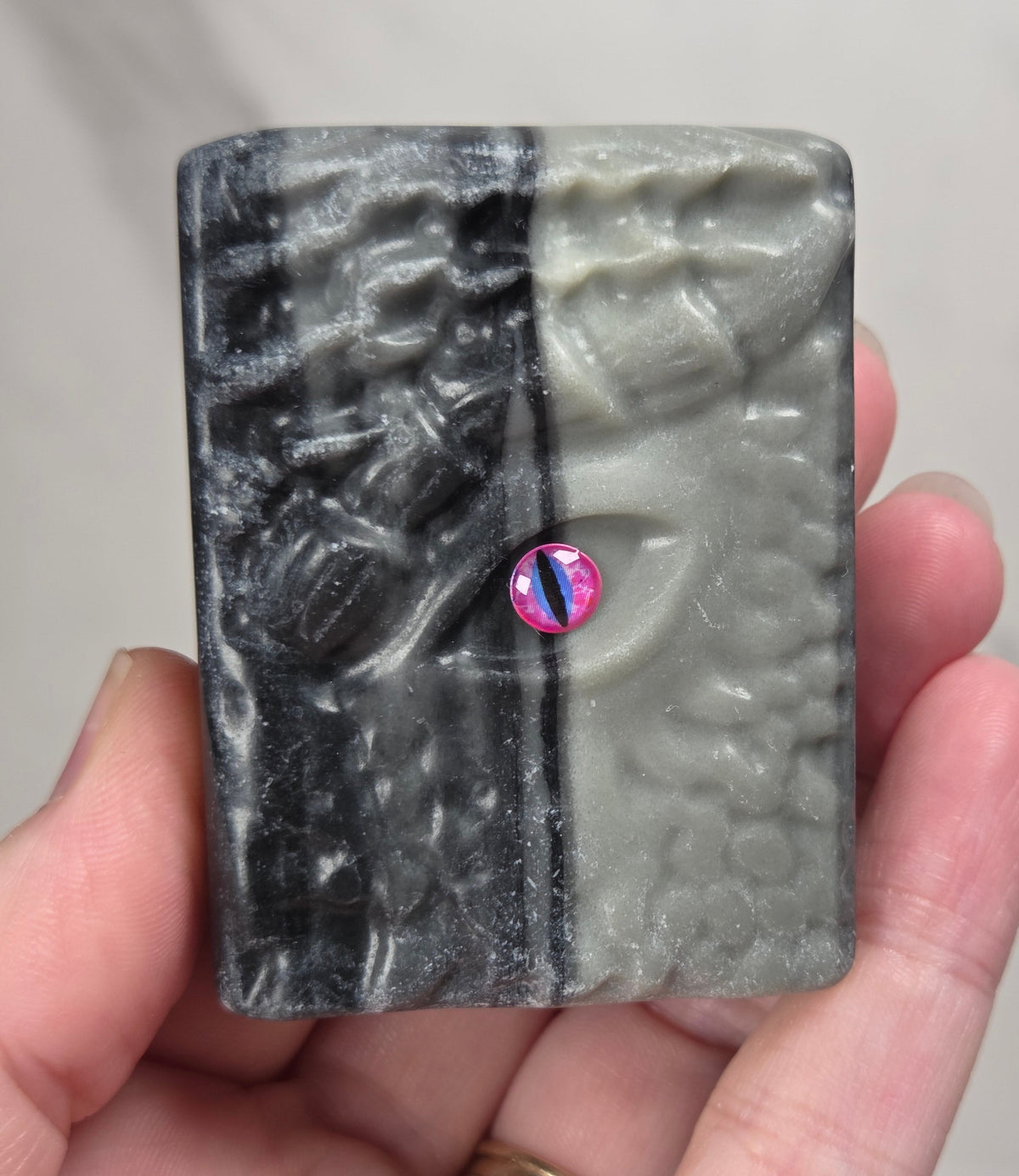 A hand holds a book carving with a split black and gray textured design and a small iridescent pink gemstone in the center, set against a light gray marble background.