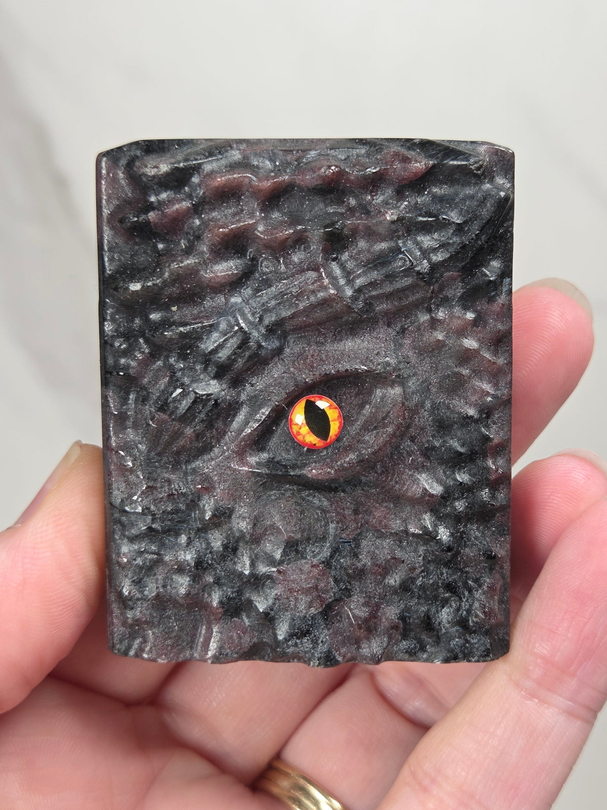 A hand holding a rectangular dark gray crystal book carving with a highly textured, reptilian or dragon-like eye design and a glowing orange-yellow pupil.