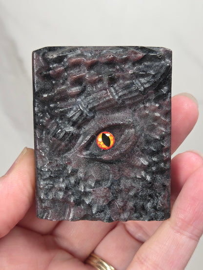 A hand holding a rectangular dark gray crystal book carving with a highly textured, reptilian or dragon-like eye design and a glowing orange-yellow pupil.