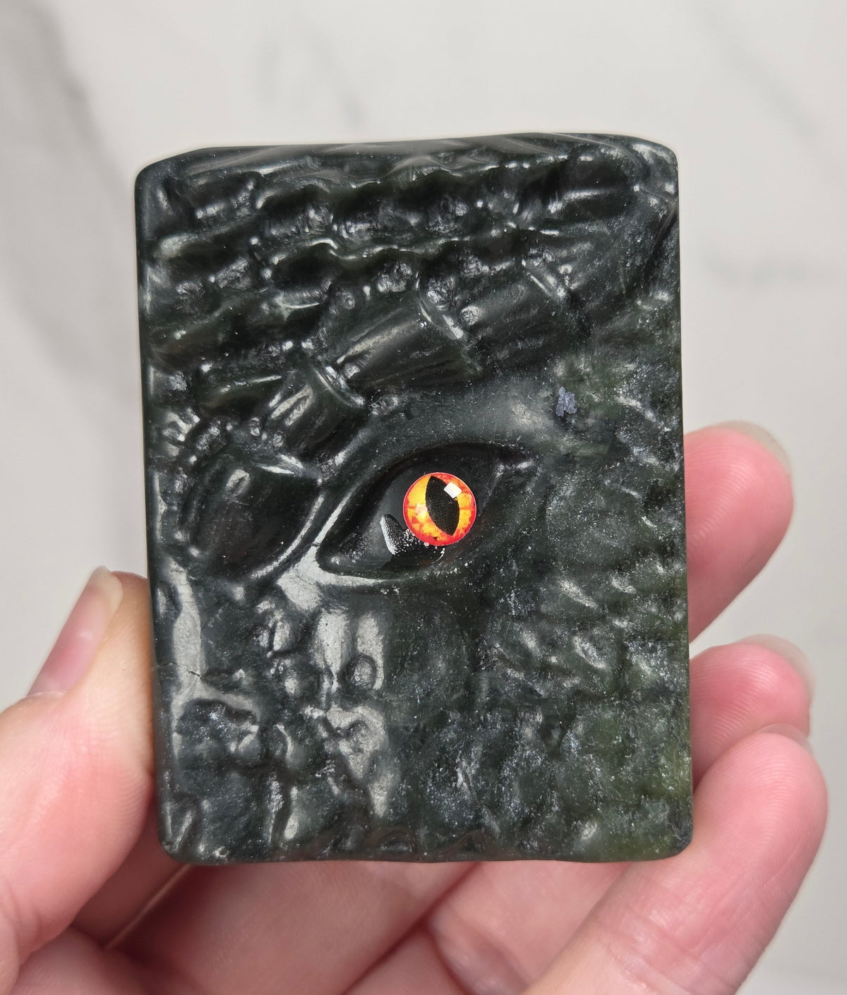A hand holding a rectangular dark green carved stone or resin plaque featuring a stylized reptile or dragon face with a glowing orange and black eye, against a light marble background.