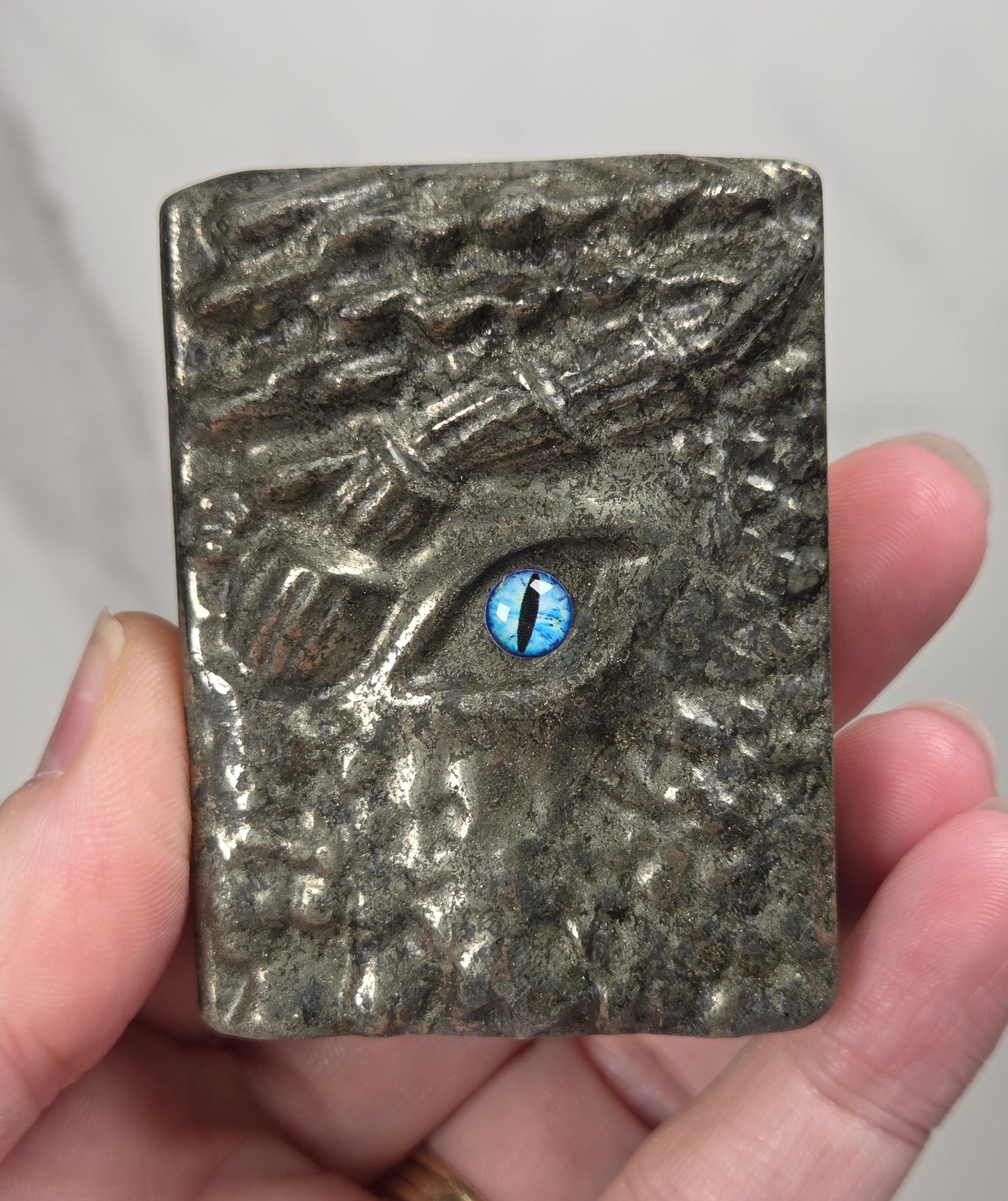 A hand holding a square metallic plaque featuring a textured animal face with a bright blue eye.