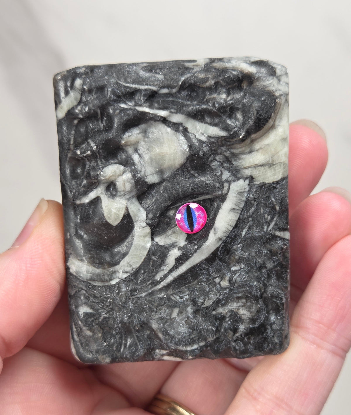 A hand holding a crystal book with a black and gray marbled pattern and a small pink and blue accent near the center, against a light gray marble background.