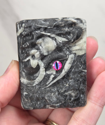 A hand holding a crystal book with a black and gray marbled pattern and a small pink and blue accent near the center, against a light gray marble background.