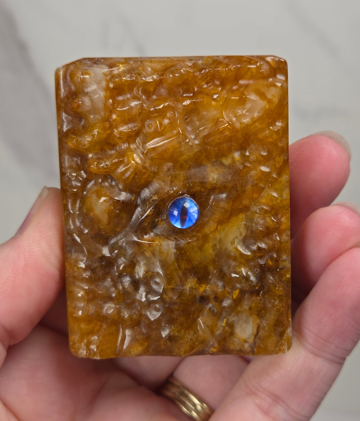A hand holding a rectangular carved amber-brown stone plaque featuring a small round blue gemstone inset near the center, against a light gray marble background.