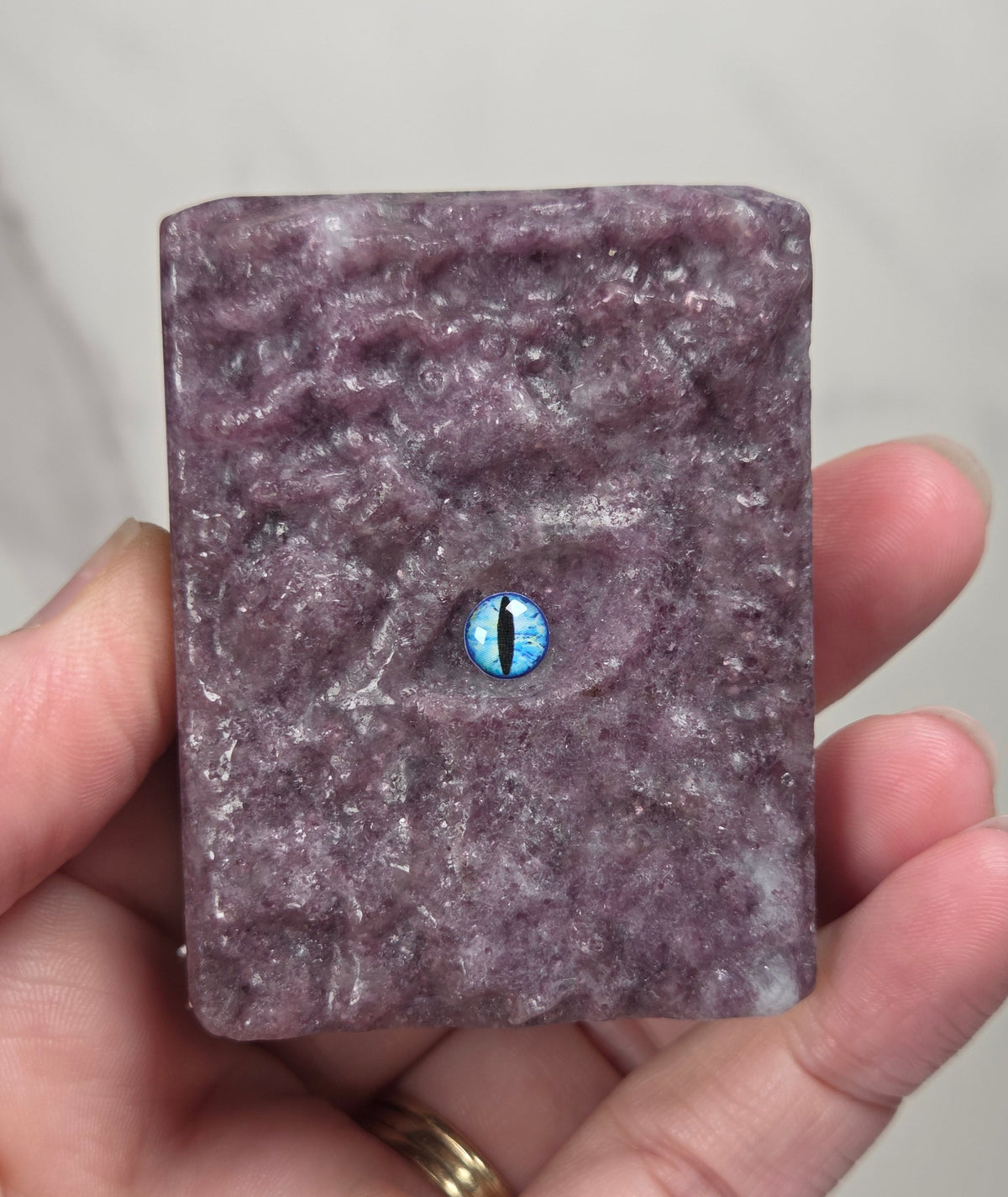 A hand holding a rectangular purple textured object with a realistic eye design in the center, featuring a blue and black iris.
