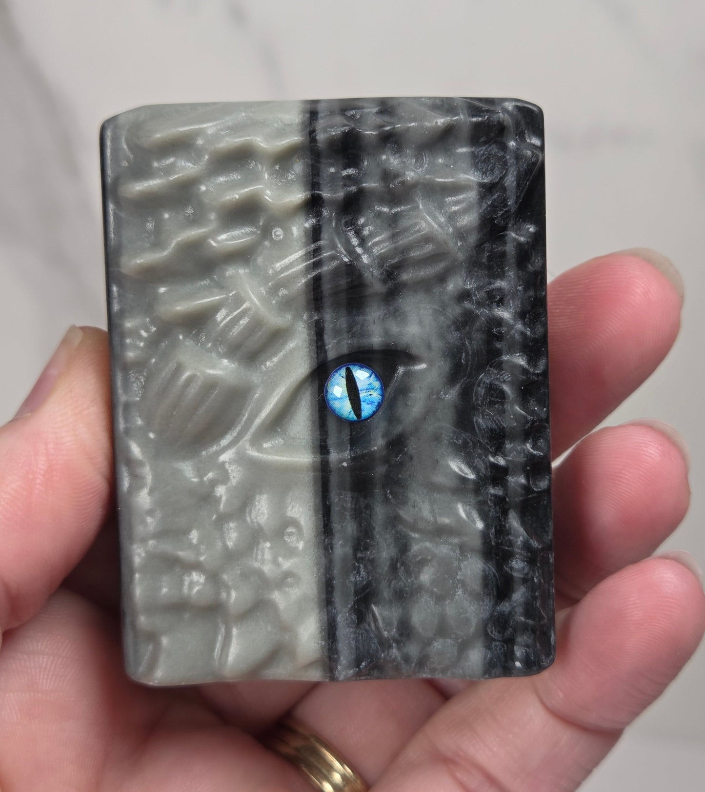 A hand holding a rectangular carved stone or resin plaque with a split design: textured light gray on the left and dark charcoal/black on the right, featuring a small blue eye motif near the center.