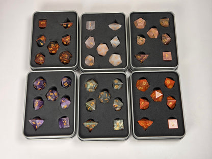 Six black metal trays containing assorted polyhedral gaming dice, arranged in a 2x3 grid on a light gray background. The trays include purple, orange, green, and brown marbled dice with gold numbers.