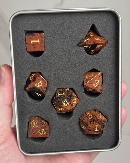 A set of seven brown wood-grain polyhedral gaming dice with gold numbers, displayed inside a gray foam-lined metal tin and held in a person's hand.