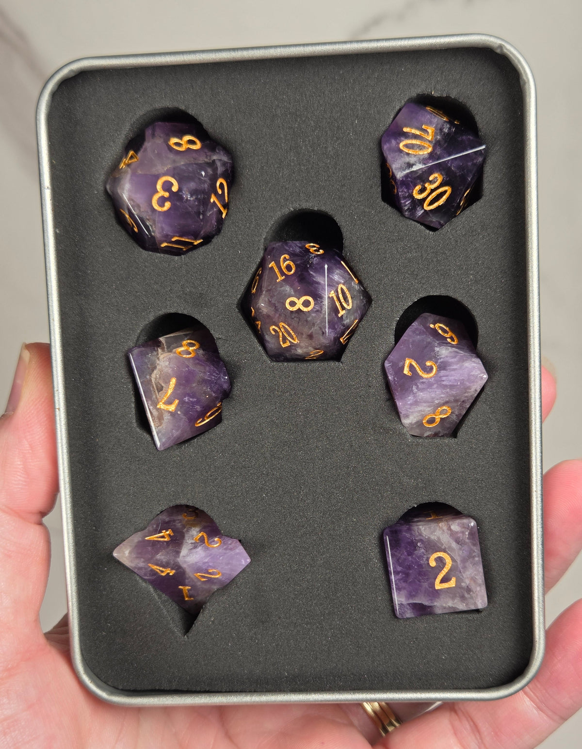 A set of purple amethyst polyhedral dice with gold-colored numbers, displayed inside a black foam-lined silver tin and held in a person's hand.