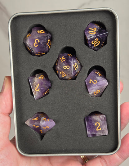 A set of purple amethyst polyhedral dice with gold-colored numbers, displayed inside a black foam-lined silver tin and held in a person's hand.