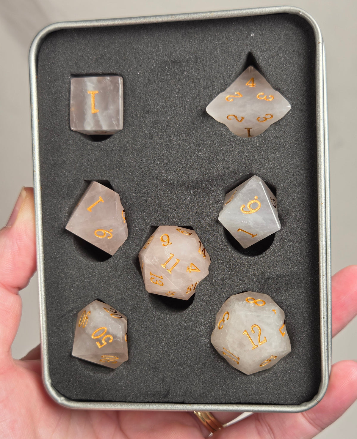 A set of translucent smoky gray polyhedral dice with gold numbers, displayed in a foam-lined metal tin held in a person's hand.