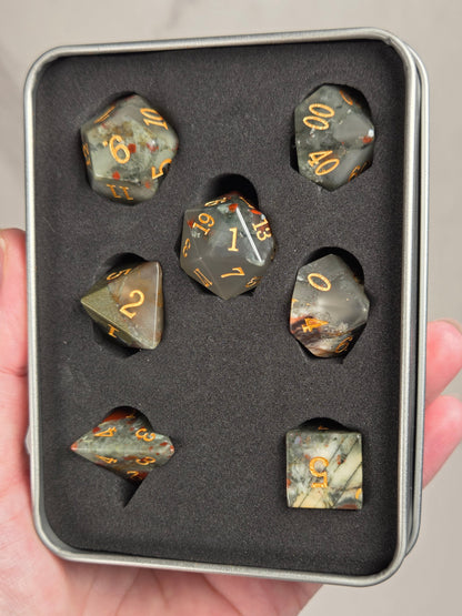 A set of seven gray polyhedral gaming dice with gold numbers, displayed inside a black foam-lined metal tin held in a person's hand.