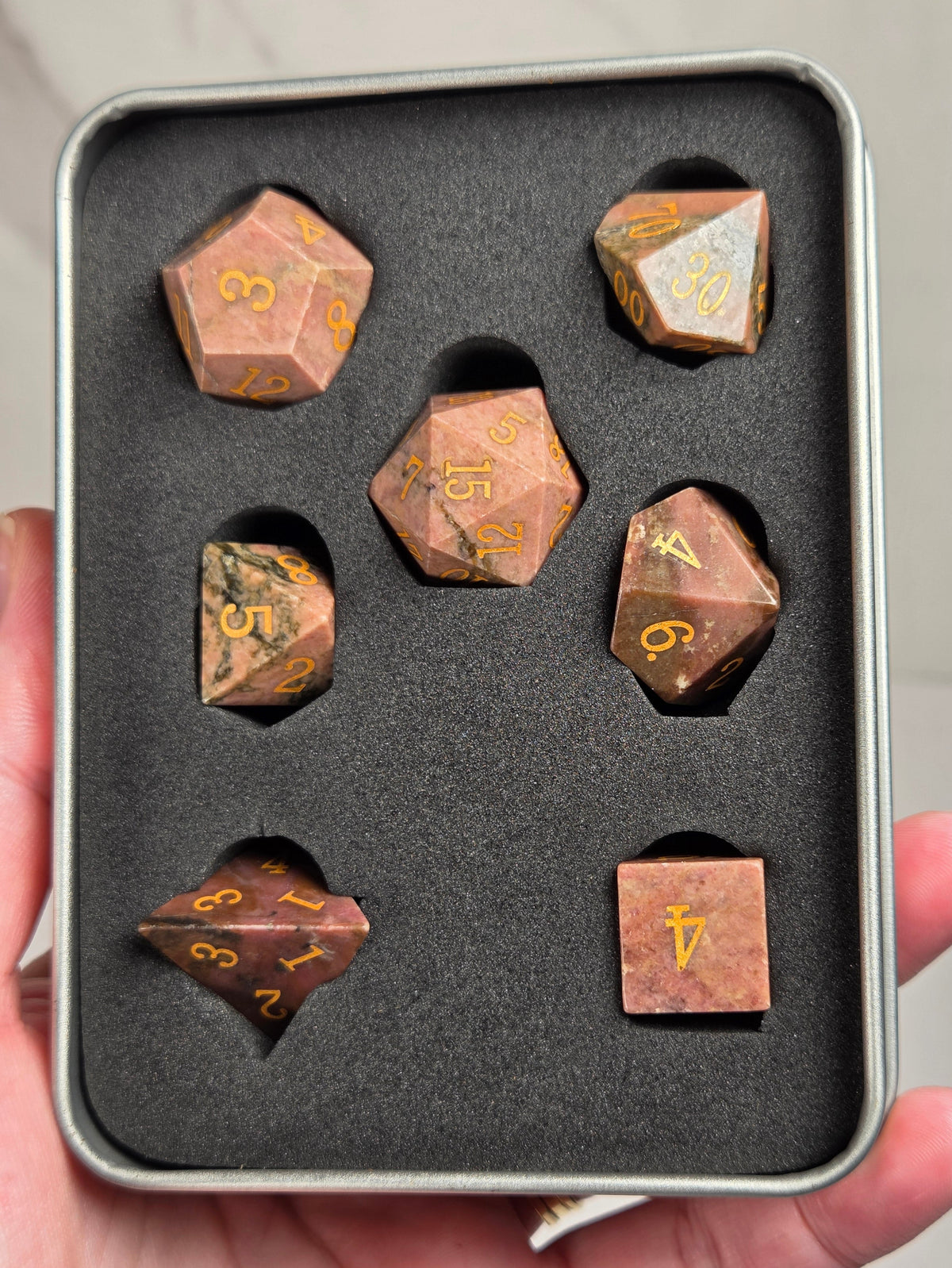 A set of polyhedral gaming dice displayed in a gray foam-lined metal tin, held in a person's hand. The dice have a coppery-brown stone texture with gold-colored engraved numbers.