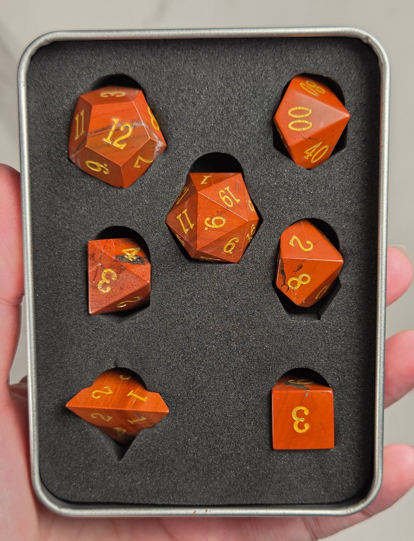 A set of orange translucent polyhedral dice with gold numbers, displayed inside a gray foam-lined metal tin held in a hand.