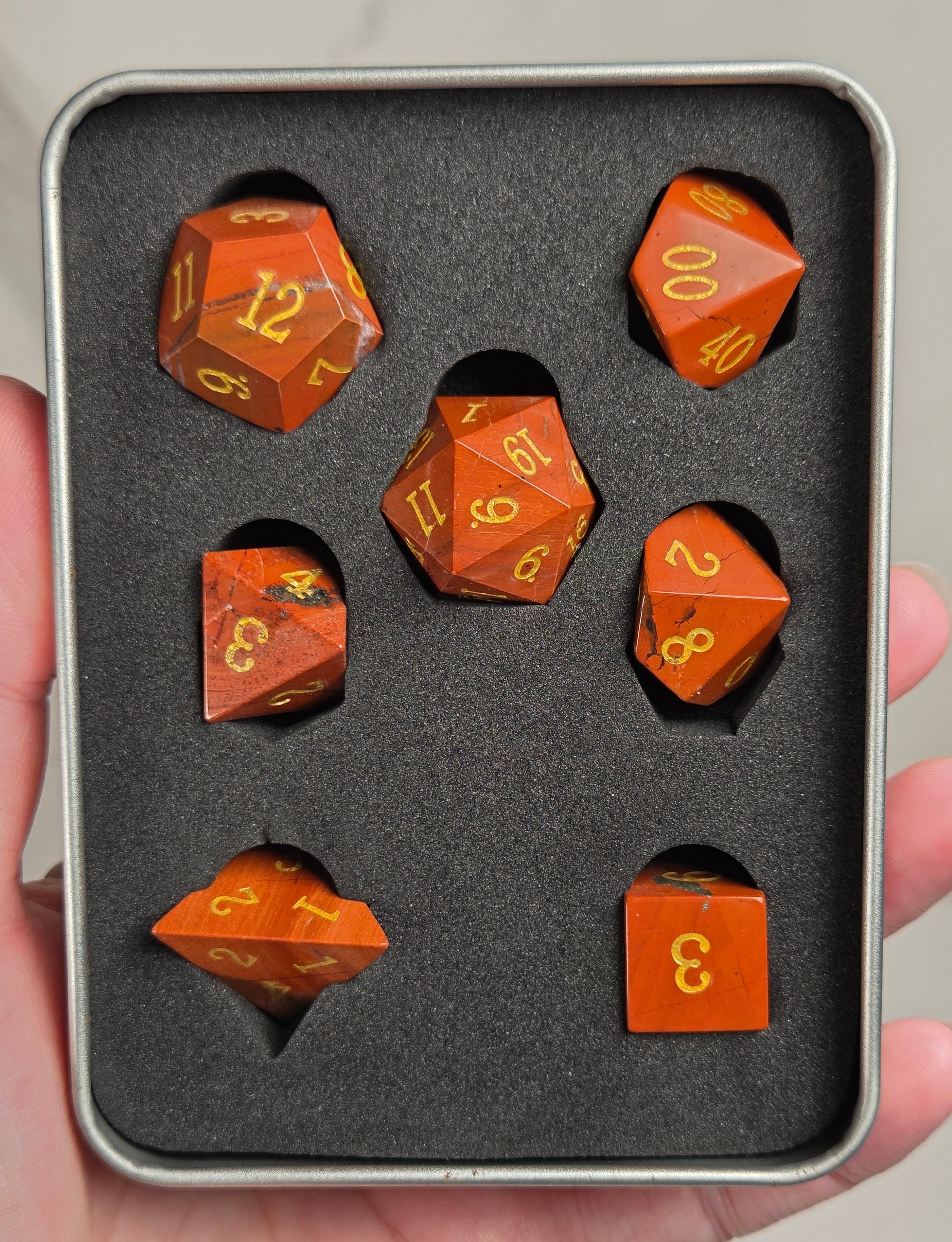 A set of orange translucent polyhedral dice with gold numbers, displayed inside a gray foam-lined metal tin held in a hand.