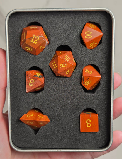 A set of orange translucent polyhedral dice with gold numbers, displayed inside a gray foam-lined metal tin held in a hand.