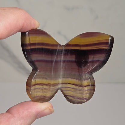 A hand holding a butterfly-shaped polished stone piece with horizontal bands of yellow, brown, purple, and dark maroon against a light gray marble background.