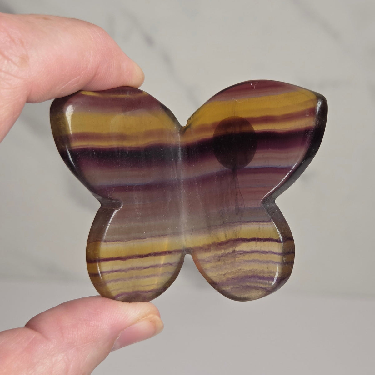 A hand holding a butterfly-shaped polished stone pendant with horizontal bands of yellow, brown, purple, and black, against a light gray marble background.